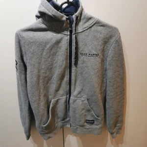 Grå hoodie från Race Marine - Säljer en grå hoodie från Race Marine med dragkedja och fickor framtill. Den har en justerbar huva med mörkblå insida och ribbade muddar vid ärmslut och nederkant. Perfekt för en avslappnad stil.