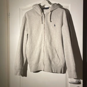 Grå hoodie från Ralph Lauren - Säljer en stilren grå hoodie från Ralph Lauren med dragkedja och huva. Den har en liten broderad logga på bröstet och praktiska fickor framtill. Perfekt för en avslappnad look. Storlek L.