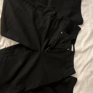 Svarta kostymbyxor från NLY Trend - Snygga svarta mid waist kostymbyxor från NLY Trend i storlek 32. Byxorna har en klassisk design med dragkedja och knapp framtill. Perfekta för en stilren look.