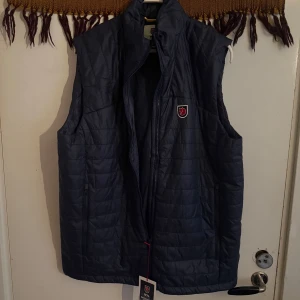 Blå quiltad väst från Fjällräven - Säljer en snygg blå quiltad väst från Fjällräven, 1974 Expedition Series. Västen har en dragkedja framtill och ett stilrent märke på bröstet. Perfekt för lager-på-lager under kyliga dagar. Passar både för äventyr och vardag.