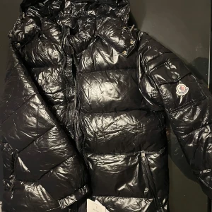Svart dunjacka från Moncler - Säljer en snygg svart dunjacka från Moncler med glansig finish. Jackan har en dragkedja framtill och en hög krage för extra värme. Perfekt för kalla dagar och har en stilren design med Moncler-logga på ärmen. Jackan har Nfc scan. A Kpia