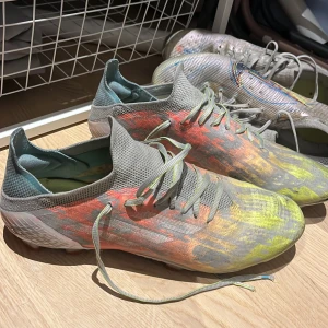 Färgglada fotbollsskor med snörning - Säljer ett par färgglada fotbollsskor med snörning. Skorna har en mix av grå, gul och orange färg med en mönstrad yta. De har en lätt och smidig design med dobbar för bra grepp på planen.