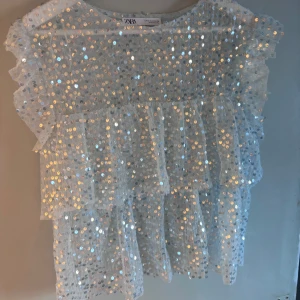 Vit/ljusblå paljettopp från Zara - Säljer en glittrig vit topp från Zara med paljetter över hela ytan. Toppen har volanger och är ärmlös, perfekt för en festlig look. Den är tillverkad i ett lätt och luftigt material. Säljer pågrund av flytt och för mycket kläder 😁😁
