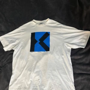 Vit t-shirt från Kenzo med blått och svart tryck - Säljer en stilren vit t-shirt från Kenzo med ett iögonfallande blått och svart grafiskt tryck på framsidan. Perfekt för dig som vill ha en unik och trendig look. T-shirten är kortärmad och tillverkad i mjukt material för extra komfort.
