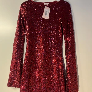 Röd glittrig klänning från Nelly - Snygg röd klänning från Nelly täckt med glittriga paljetter. Den har långa ärmar och en figurnära passform som gör den perfekt för festliga tillfällen. Klänningen är i en mörkröd nyans som verkligen sticker ut. Helt oanvänd med ett nypris på 600kr 