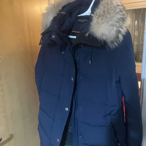 Mörkblå dunjacka från Hollies - Snygg mörkblå vinterjacka från Hollies med pälskantad huva. Jackan har dragkedja och knappar framtill samt långa ärmar. Perfekt för kalla dagar.