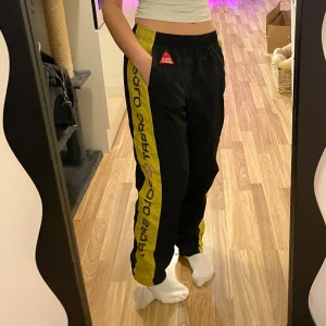 Svarta träningsbyxor från Polo Sport - Snygga svarta träningsbyxor från Polo Sport med gula detaljer och text längs benen. Byxorna har en elastisk midja och en röd Polo Sport-logga på sidan. Perfekta för en sportig look. Jag är 160cm lång 