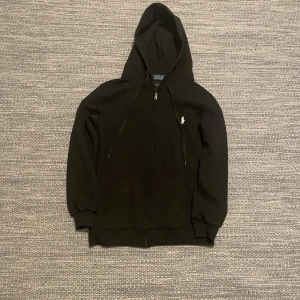 Svart zip hoodie från Ralph Lauren (Aldrig använd) - Säljer en stilren svart hoodie från Ralph Lauren med dragkedja och huva. Den har långa ärmar och en liten broderad logga på bröstet. Perfekt för en avslappnad look. 