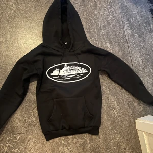 Svart corteiz hoodie  - Säljer en svart hoodie med ett coolt tryck på framsidan. Den har en klassisk känguruficka och justerbar huva med snören. Perfekt för en avslappnad stil.