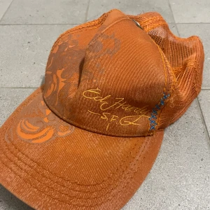 Orange keps från Ed Hardy - Säljer min keps från Ed Hardy med meshdetaljer och ett unikt tryck. Kepsen har blå sömmar och en metallknapp med dödskallemotiv. 