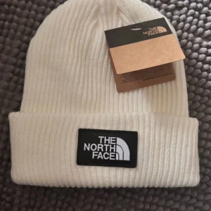 Vit beanie från The North Face - Snygg vit ribbad mössa från The North Face med klassisk logga framtill. Perfekt för kyliga dagar och håller dig varm med stil. Passar både till vardags och utomhusaktiviteter.
