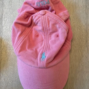 Rosa keps från Polo Ralph Lauren - Snygg rosa keps från Polo Ralph Lauren med broderad logga i blått framtill. Kepsen har en justerbar rem baktill för perfekt passform. Perfekt för en avslappnad stil.
