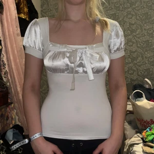 Vit satinblus med rosett - Elegant vit blus i satin med korta puffärmar och en söt rosett framtill. Blusen har en smickrande passform och kan bäras både med och utan axlar. Perfekt för en stilfull look.