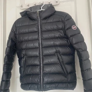 Svart dunjacka från Colmar - Säljer en stilren svart dunjacka från Colmar. Jackan har en dragkedja framtill och två praktiska fickor med dragkedjor. Perfekt för kyliga dagar med sin vadderade design och höga krage. Dock har den vita små prickar på sig med inget man tänker på