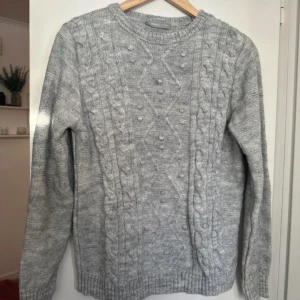 Grå stickad tröja från lc Waikiki  - Säljer en mysig grå stickad tröja från lc Waikiki med flätmönster och små bollar på framsidan. Tröjan har långa ärmar och ribbstickade muddar vid ärmslut och nederkant. Perfekt för kyliga dagar! 🧶
