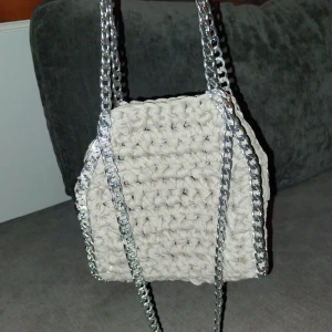 Beige virkad väska med silverkedjor - Säljer en unik beige virkadväska med coola silverkedjor som handtag. Perfekt för att ge din outfit en trendig touch. Väskan har en fyrkantig form och är handgjord med ett grovt stickat mönster. Passar perfekt för dig som vill ha något annorlunda och stilrent.