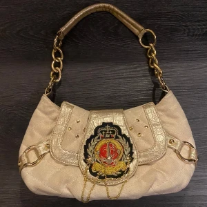 Y2K Guldig axelväska med broderad detalj  - Snygg beige axelväska med guldfärgade kvalitativ  riktig metall kedjehandtag och en unik broderad emblem på framsidan. Väskan har snygg 2000 stil design och guld glitter detaljer för extra glam. Perfekt för en Y2K baddie. 😘