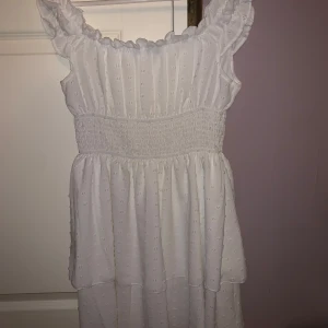 Vit klänning från Shein - Söt vit klänning från Shein med volangdetaljer och smockad midja. Klänningen är ärmlös och har en luftig design som passar perfekt för varma dagar. Den har ett diskret prickigt mönster som ger en extra touch av stil.