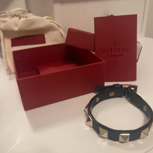 Blå Valentino armband - Snyggt blå armband från Valentino med guldiga  nitar. Armbandet kommer i sin originalförpackning med en röd låda och tygpåse. Perfekt accessoar för att ge en edgy touch till din outfit samt fester.