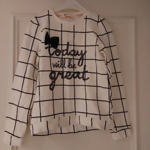 Vit rutmönstrad barn tröja med text,storlek 10 år - Säljer en vit tröja till flickor från H&M med svart rutmönster och texten 'today will be great' i pärlor. Tröjan har en dekorativ svart rosett på bröstet och långa ärmar. Perfekt för en stilren och trendig look.