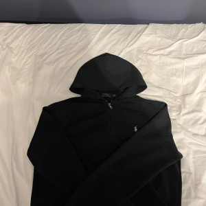 Svart hoodie från Ralph Lauren - Säljer en stilren svart hoodie från Ralph Lauren med dragkedja framtill. Tröjan har en klassisk design med huva och långa ärmar
