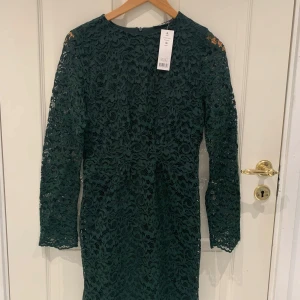 Grön spetsklänning - Säljer en elegant grön spetsklänning med långärmad design. Klänningen har ett vackert blommigt mönster och är perfekt för festliga tillfällen. Den är helt nytt  och har en normal passform.