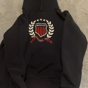 Mörk blå hoodie från GANT - Säljer en mörk blå hoodie från GANT med ett stort emblem på framsidan. Hoodien har en klassisk design med känguruficka och ribbade muddar. Perfekt för en avslappnad stil.