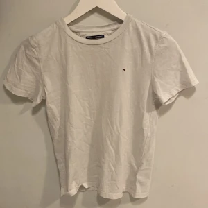 Vit t-shirt från Tommy Hilfiger - Säljer en klassisk vit t-shirt från Tommy Hilfiger med en liten broderad logga på bröstet. T-shirten är tillverkad i 100% bomull och har en rund halsringning. Perfekt för en stilren och avslappnad look.