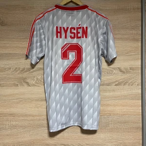 Liverpool Borta Hysén 2 L - Säljer en grå fotbollströja från Adidas med röd text och nummer 2 på ryggen. Tröjan har korta ärmar och ett mönster i grått. Framsidan pryds av loggan för Liverpool FC och texten 'Candy'. Perfekt för samlare eller fans!
