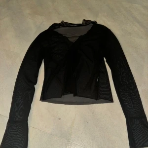 Svart mesh topp - Snygg svart mesh topp med långärmad design. Perfekt för en kväll ute eller att bära över ett linne för en lager-på-lager-look. Toppen har en omlottstil framtill som ger en trendig touch.