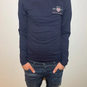 Mörkblå långärmad tröja från GANT - Säljer en stilren mörkblå långärmad tröja från GANT med logotyp och text på bröstet. Perfekt för en avslappnad och snygg look. Passar bra till jeans eller chinos.