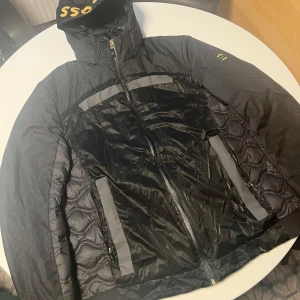 Svart jacka från BOSS - Snygg svart jacka från BOSS med guld logga och detaljer. Jackan har en quiltad design och dragkedja framtill. Perfekt för kyligare dagar med sin stilrena look.
