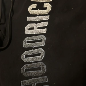 Svart hoodie från Hoodrich - Säljer en svart hoodie från Hoodrich med stor logga i vitt till grått på framsidan. Den har en klassisk design med dragsko i huvan och en stor magficka. 