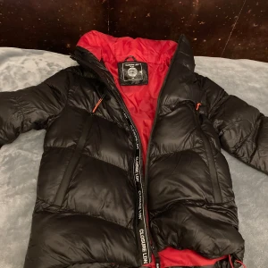 Svart dunjacka från Closure LDN - Säljer en snygg svart dunjacka från Closure LDN med röd insida. Jackan har en praktisk dragkedja och långa ärmar. Perfekt för kyliga dagar med sin pufferdesign och huva. Använd endast ett fåtal gånger: