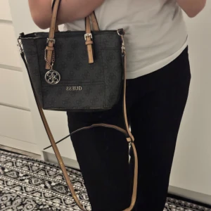 Svart axelväska från Guess - Snygg svart axelväska från Guess med mönstrad yta och justerbara remmar i beige. Väskan har en praktisk dragkedja och en dekorativ metallberlock. Perfekt för att bära dina dagliga nödvändigheter med stil.