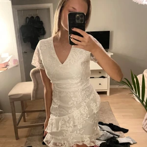 Vit spetsklänning med volangdetaljer - Elegant vit klänning med v-ringning och korta ärmar. Klänningen har vackra spetsdetaljer och en volang som ger en romantisk känsla. Perfekt för sommarens festligheter eller studenten 