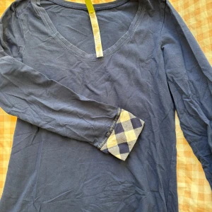Blå långärmad tröja / pyjamas  - Säljer en blå långärmad tröja från Hue med v-ringning. Tröjan har ett rutigt mönster i blått och beige vid ärmsluten som ger en snygg detalj. Perfekt för en avslappnad stil.