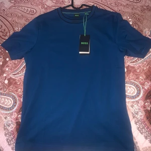 Blå t-shirt från Boss - Snygg blå t-shirt från Boss i ett klassiskt snitt. Perfekt för en stilren look. T-shirten har korta ärmar och är tillverkad i ett mjukt material.