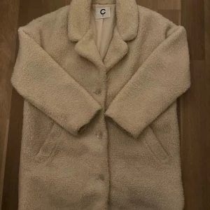Beige teddyjacka från C - Mysig beige teddyjacka från C i storlek M. Jackan har en klassisk krage och knäppning framtill. Perfekt för kyliga dagar med sin mjuka och varma textur.