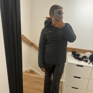 Peak performance jacka  - Säljer en svart dunjacka med dragkedja och huva. Jackan har en quiltad design och är perfekt för kyligare väder. Den har en normal passform och långa ärmar. Ett hål i fickan men inget som syns