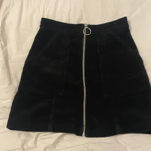 Svart manchesterkjol  - Snygg svart manchesterkjol i storlek XS. Kjolen har en dragkedja framtill som ger en cool detalj. Perfekt för att skapa en trendig look. Använd några gånger