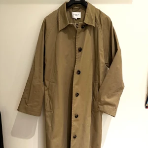Ny beige trenchcoat från Stockh LM Studio - Snygg beige trenchcoat från Stockh LM Studio i storlek M. Helt ny aldrig använd men som har ett litet solblekt område på ena sidan. Jackan har en klassisk design med knappar framtill och en stilren krage. Perfekt för vår och höst. Har en oversized passform och täcker nästan hela benlängden (är 165cm lång). Fler bilder kan skickas! 