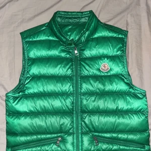 Grön dunväst från Moncler - Snygg grön dunväst från Moncler med dragkedja och två fickor framtill. Västen har en quiltad design och Monclers logotyp på bröstet. Perfekt för kyligare dagar.