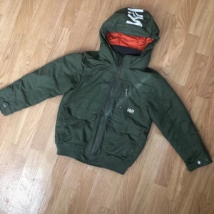 WARP Jacka  - Säljer en grön jacka med huva från WA. Jackan har en dragkedja framtill och en praktisk bröstficka. Insidan är fodrad med orange tyg för extra värme. Perfekt för kyliga dagar!