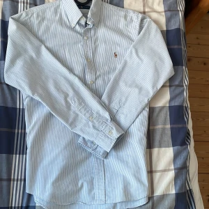 Blå- och vitrandig skjorta från Ralph Lauren - Säljer en klassisk blå- och vitrandig skjorta från Ralph Lauren. Skjortan har långa ärmar och knappar framtill. Perfekt för en stilren look.