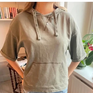 Olivgrön hoodie med korta ärmar - Säljer en olivgrön hoodie med korta ärmar och en praktisk magficka. Den har en avslappnad passform och justerbar huva med snören. Perfekt för en casual look!
