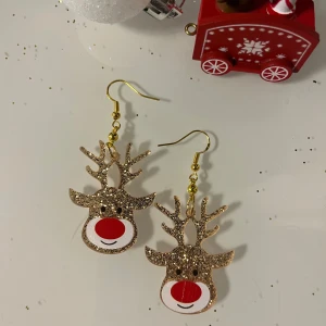 Glittriga renörhängen - Charmiga örhängen i form av renar med glittrig yta och röd nos. Perfekta för att sprida julstämning med sin lekfulla design. De hängande örhängena har guldfärgade krokar och detaljer.