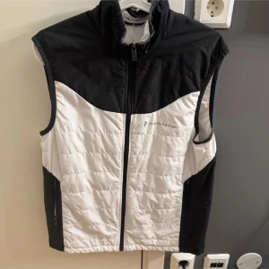 Svart och vit dunväst från Peak Performance - Snygg och funktionell svart och vit dunväst från Peak Performance. Västen har en dragkedja framtill och är perfekt för kyligare dagar. Den är ärmlös och har en stilren design med logotyp på bröstet.