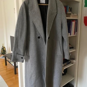 Grå kappa - Snygg grå kappa med dubbelknäppning och breda slag. Den har långa ärmar och två fickor framtill. Perfekt för kyligare dagar.