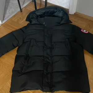 Canada goose jacka - Säljer en svart dunjacka från Canada Goose i storlek M. Jackan är perfekt för kalla vinterdagar och har en klassisk design med huva och knappar. Den är i mycket bra skick och håller dig varm och bekväm. Passar både till vardags och mer formella tillfällen.